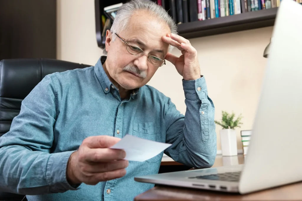 An Elderly Man Reading Paperwork with a Laptop 