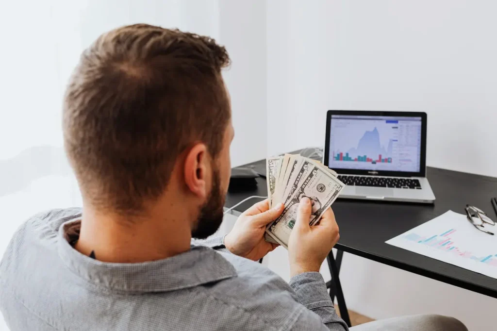 A Man Counting Money His Money at a Desk with a Laptop 